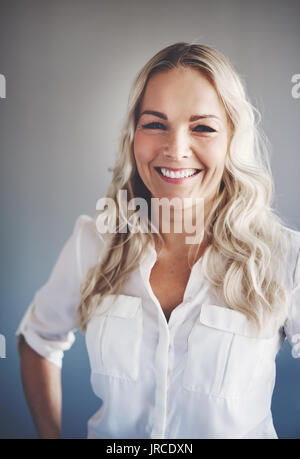 Portrait of a smiling young businesswoman with blonde hair standing alone in an office with her hands on her hips Stock Photo