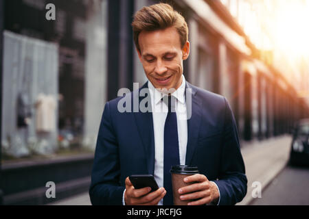 Smiling young businessman reading text messages on his cellphone and drinking a coffee while standing on a city street in the morning Stock Photo
