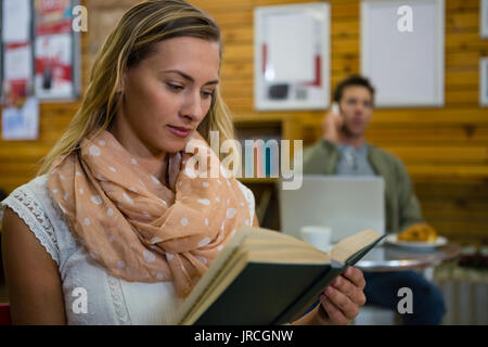 Woman reading book while man talking on phone in background at cafe Stock Photo