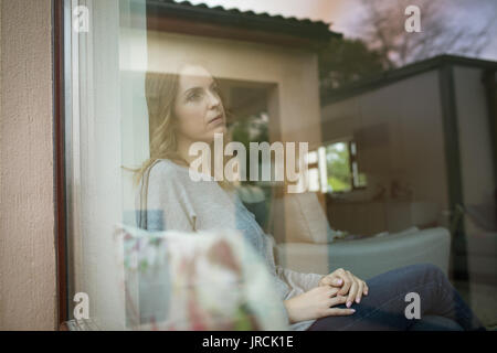 Thoughtful woman sitting by window seen through glass Stock Photo