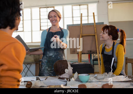 Smiling teacher holding laptop while talking to students sitting in art class Stock Photo