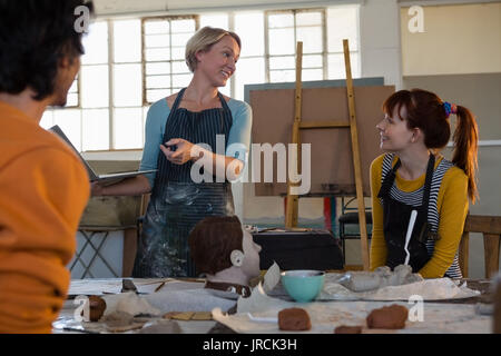 Female teacher holding laptop while talking to students sitting in art class Stock Photo