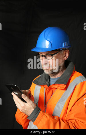 A construction engineer in an orange high-viz jacket studies a tablet computer. Plain, dark background. Stock Photo