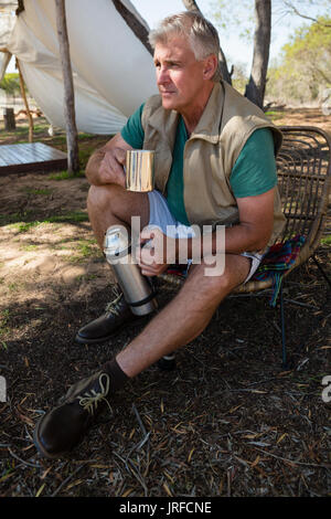 Mature man holding coffee cup and thermos while sitting on chair at campsite Stock Photo