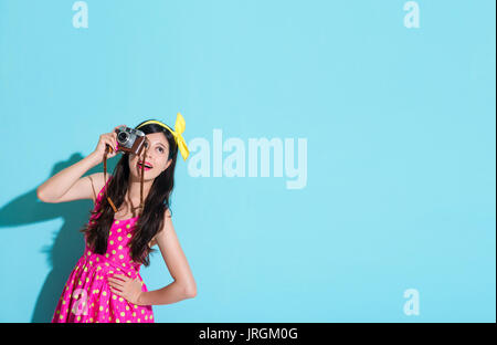 happy beautiful woman using vintage style camera standing in blue background and wearing summer dress looking at empty area feeling cheerful. Stock Photo