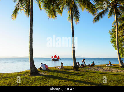 A sunset cruise Chinese junk sails past people relaxing in Rex Smeal Park, Port Douglas, Far North Queensland, FNQ, QLD, Australia Stock Photo