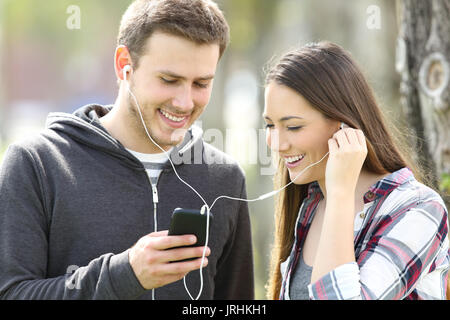 Happy couple of teenagers sharing on line music and watching media content in a smart phone outdoors in a park Stock Photo