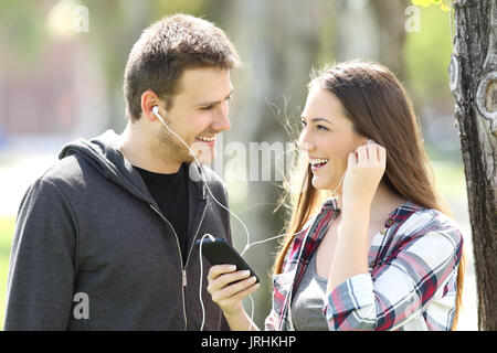 Happy couple of teens sharing on line music in a smart phone outdoors in a park Stock Photo