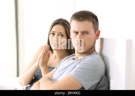 Wife begging to her angry husband sitting on the bed in a house interior Stock Photo