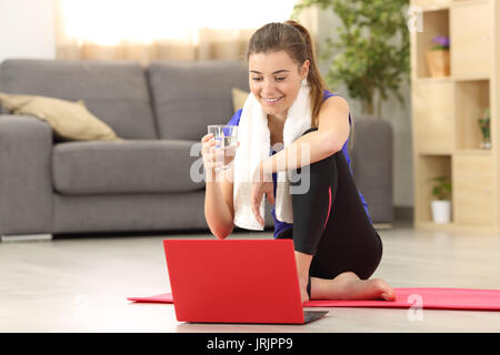 Front view of a fitness woman holding a water glass watching on line tutorials sitting on the floor in the living room at home Stock Photo