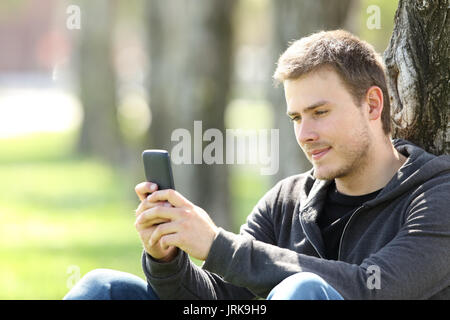 Portrait of a single boy reading messages in a smart phone sitting outdoors in a park Stock Photo