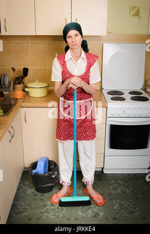 A depressed  adult woman, a housewife or a maid, wearing a red apron and a green scarf on her head is resting after she has swept the kitchen with a b Stock Photo