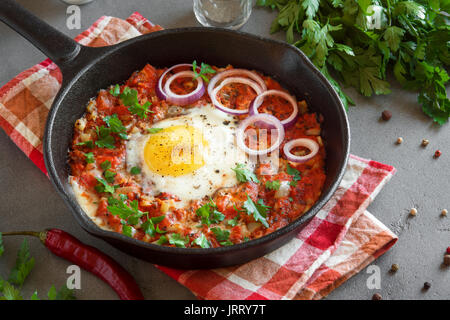 Shakshuka in a Cast Iron Pan. Middle eastern style eggs poached in tomato and pepper sauce with parsley, onion and spices on grey concrete background. Stock Photo
