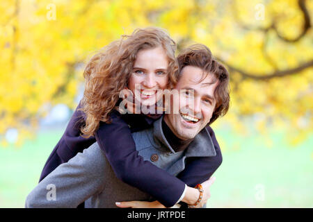 Love, relationships, season and people concept - happy young couple having fun in autumn park piggyback ride Stock Photo