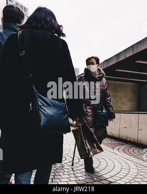 Japanese man walks down the street with a mask on his face as two people walk past him. Stock Photo