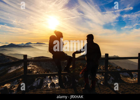 Silhouette of young woman and man with backpacks and sticks on mountains at sunset. Stock Photo
