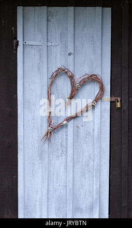 Old wooden shed painted in black and blue color. The door is decorated with a heart made of sticks and flowers. Stock Photo