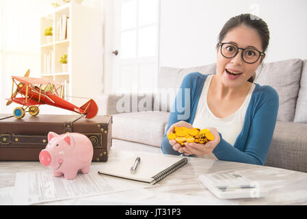 happy asian woman showing smile hands holding gold money coins from piggybank saving sitting on sofa in the living room at home. travel and vacation f Stock Photo