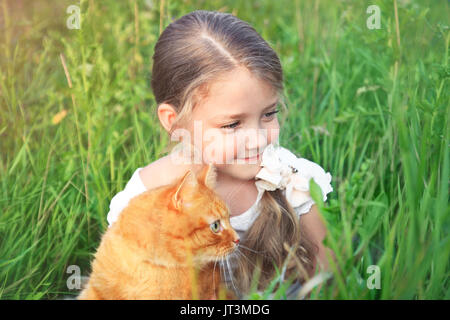 Cute little girl is holding a red cat in nature sitting in the grass. Stock Photo