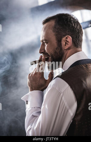 Side view of smiling confident man smoking cigar indoors  Stock Photo