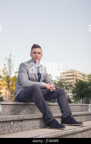 one young Caucasian man, blank expression, business suit, formal wear, ordinary common person portrait, outdoors sitting on stairs steps Stock Photo