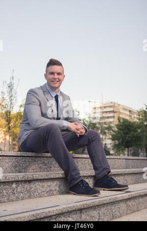 one young Caucasian man smiling, business suit, formal wear, ordinary common person portrait, outdoors sitting on stairs steps Stock Photo