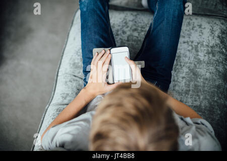 Shot from above of a boy sitting on his sofa at home, using a smartphone. Stock Photo