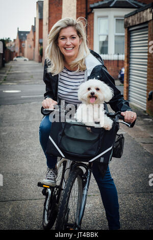 Mature woman sitting on her bike with her pet Bichon Frise in a basket on the front. They are both looking at the camera; the woman is smiling and the Stock Photo