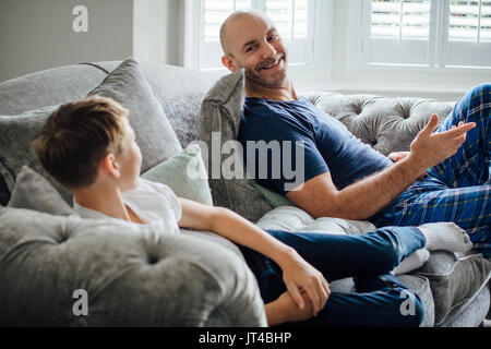Father and son sitting on the sofa in their home together in teh morning. They are talking and laughing about something they are watching on televisio Stock Photo