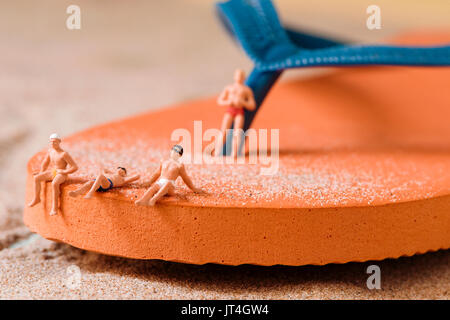 some different miniature men wearing swimsuit relaxing on an orange flip-flop on the sand of the beach Stock Photo
