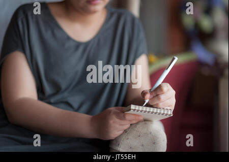 Left-handed woman writing some idea on small notepad while sitting on chair in house. Freelance working from anywhere concept Stock Photo