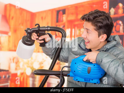 Close up of a handsome smiling man holding in his hands the painting spray gun, an wearing a gray jacket in a blurred background, profile view Stock Photo