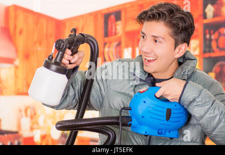 Close up of handsome smiling man holding in his hands the painting spray gun, an wearing a gray jacket in a blurred background Stock Photo