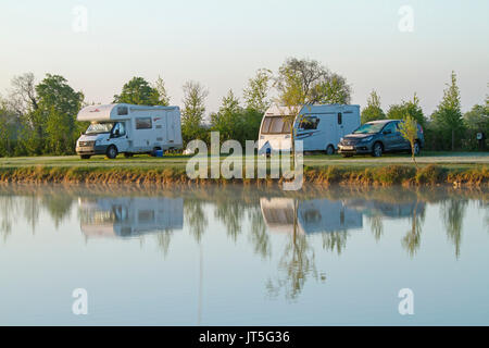 Caravan and motorhome beside trees in rural landscape reflected in calm blue water of coarse fishing pond under blue sky in England Stock Photo