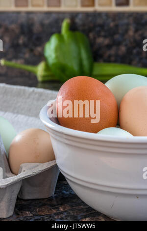 Vertical photo of multicolored eggs in a white bowl and some in the carton to the side on a kitchen counter with a green pepper and okra in the backgr Stock Photo