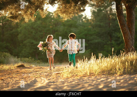 Little girl with bouquet and boy are running in sunny forest road and holding hands. First love. Stock Photo