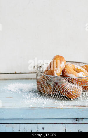 Homemade donuts with sugar powder in frying basket served with vintage sieve on blue wooden table. Rustic style, day light. Stock Photo