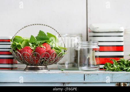 Fresh ripe garden strawberries and melissa herbs in vintage vase standing with empty glass and metal jars for jam on blue white wooden kitchen table.