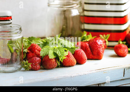 Fresh ripe garden strawberries and melissa herbs with empty glass and vintage metal jars for jam on blue white wooden kitchen table. Rustic style, day Stock Photo