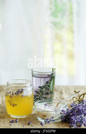 Organic raw honey, white sugar in glass jars, glass of water flavored with lavender flowers, standing on table with sackcloth. Rustic style, day light Stock Photo