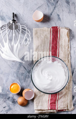 Process of cooking meringue. Whipped egg whites on mixer whisk with broken eggs on linen towel over gray texture background. Top view. Baking dessert Stock Photo