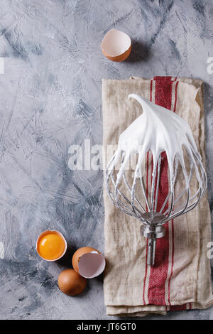 Process of cooking meringue. Whipped egg whites on mixer whisk with broken eggs on linen towel over gray texture background. Top view. Baking dessert Stock Photo