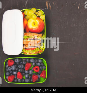 Sandwich, apple, grapes, carrot, berry in plastic lunch box on black chalkboard. Back to school concept. Stock Photo