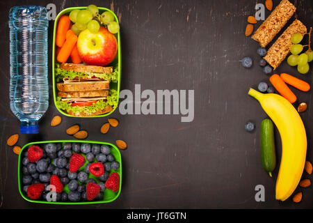 Sandwich, apple, grape, carrot, berry in plastic lunch box and bottle of water on black chalkboard. Back to school concept. Stock Photo