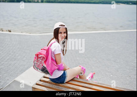 Fantastic young woman in casual clothing and cap sitting on the bench in the skatepark with rollerblades on. Stock Photo