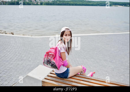 Fantastic young woman in casual clothing and cap sitting on the bench in the skatepark with rollerblades on. Stock Photo