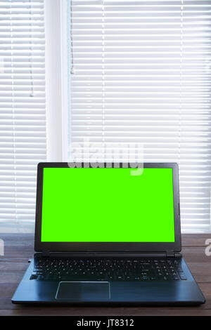 laptop in small home office on wooden desk with green screen on display and lot of copy space. vertical shot Stock Photo