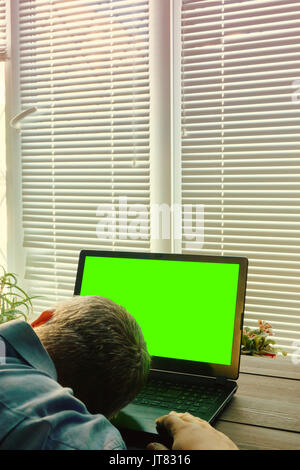 concept image of overtime work. tired man fall asleep while working on a laptop till sunrise. mans head laying on a laptop keyboard. laptop with green Stock Photo