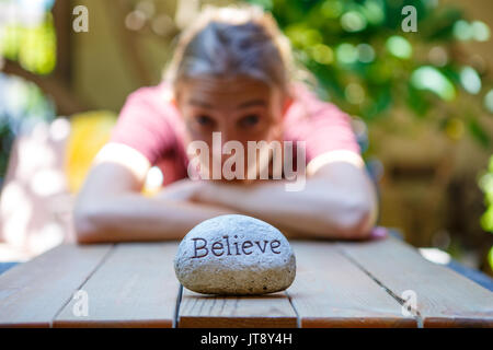 Young woman sitting at a wooden table in the background with a rock in front of her inscribed with the word believe .model release Stock Photo