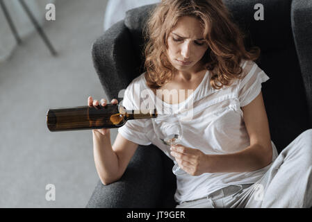 Alcohol addiction. Upset moody cheerless woman sitting in the armchair and drinking wine while having problems with alcohol Stock Photo